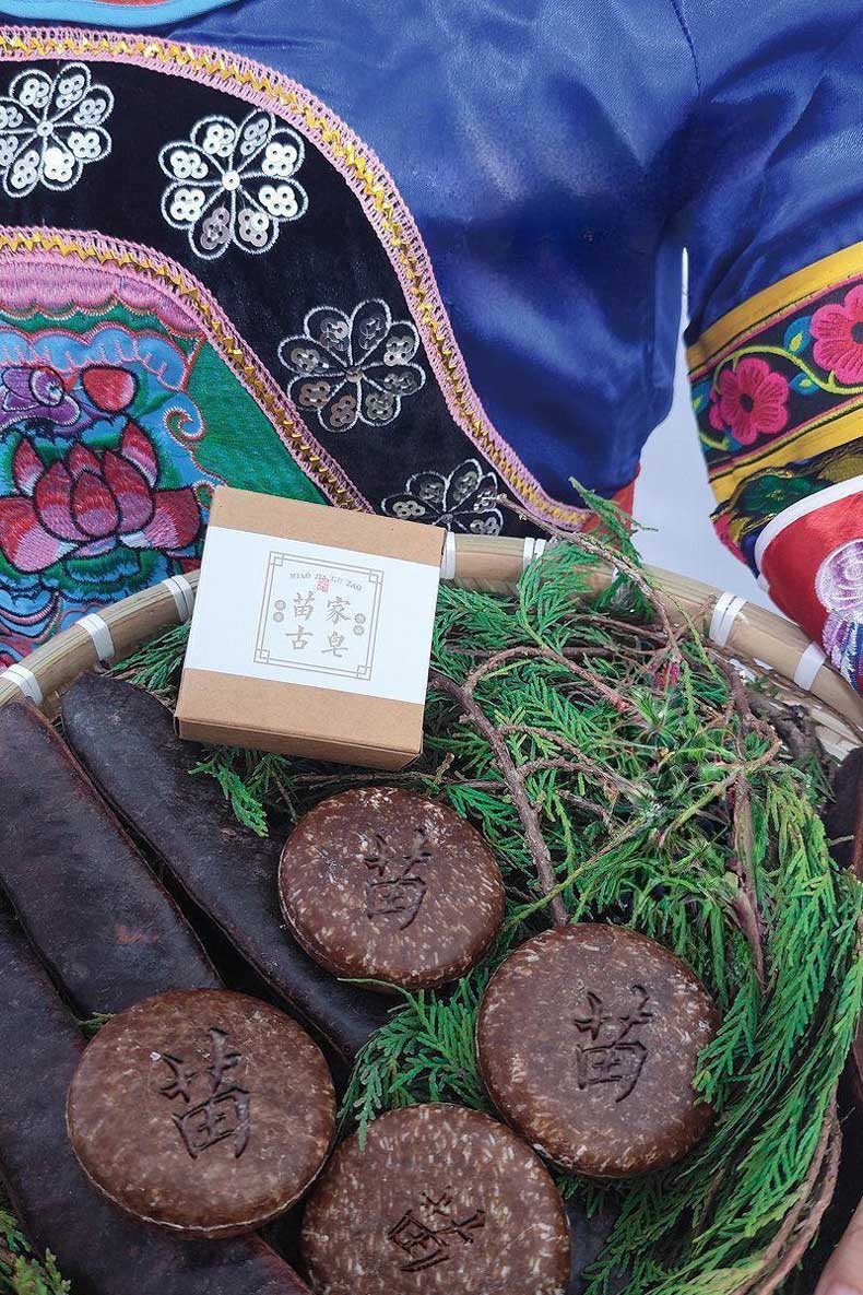A display of Miao - family herbal soaps in a bamboo basket, with a box labeled "Miao Family Ancient Soap" and surrounded by natural plants. The person holding the basket is wearing traditional Miao - ethnic - style clothing.