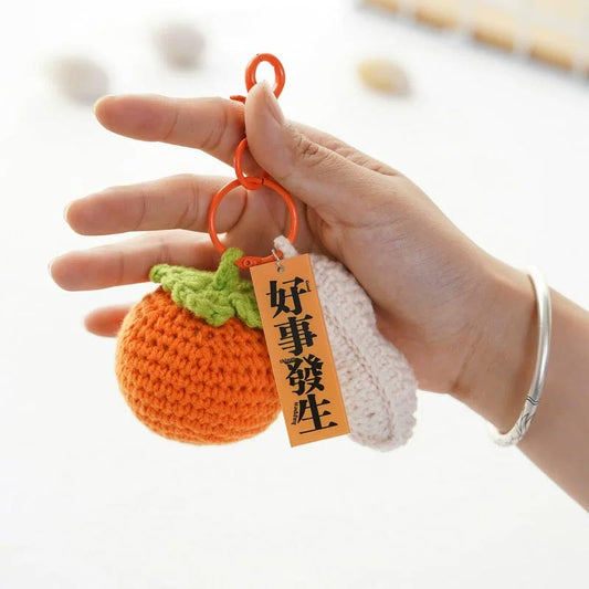 A hand gracefully holds a charming keychain, crafted from intricately knitted orange and white threads, shaped like a festive pumpkin adorned with a verdant topper. Dangling from it, an orange tag proclaims, "Good Things Will Happen," spreading a message of positivity and good fortune wherever it goes.